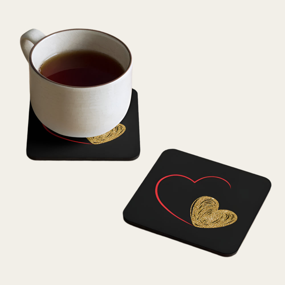 Two square coasters with a cork backing, with a red and gold heart print, displayed with a cup of tea on one of the coasters.